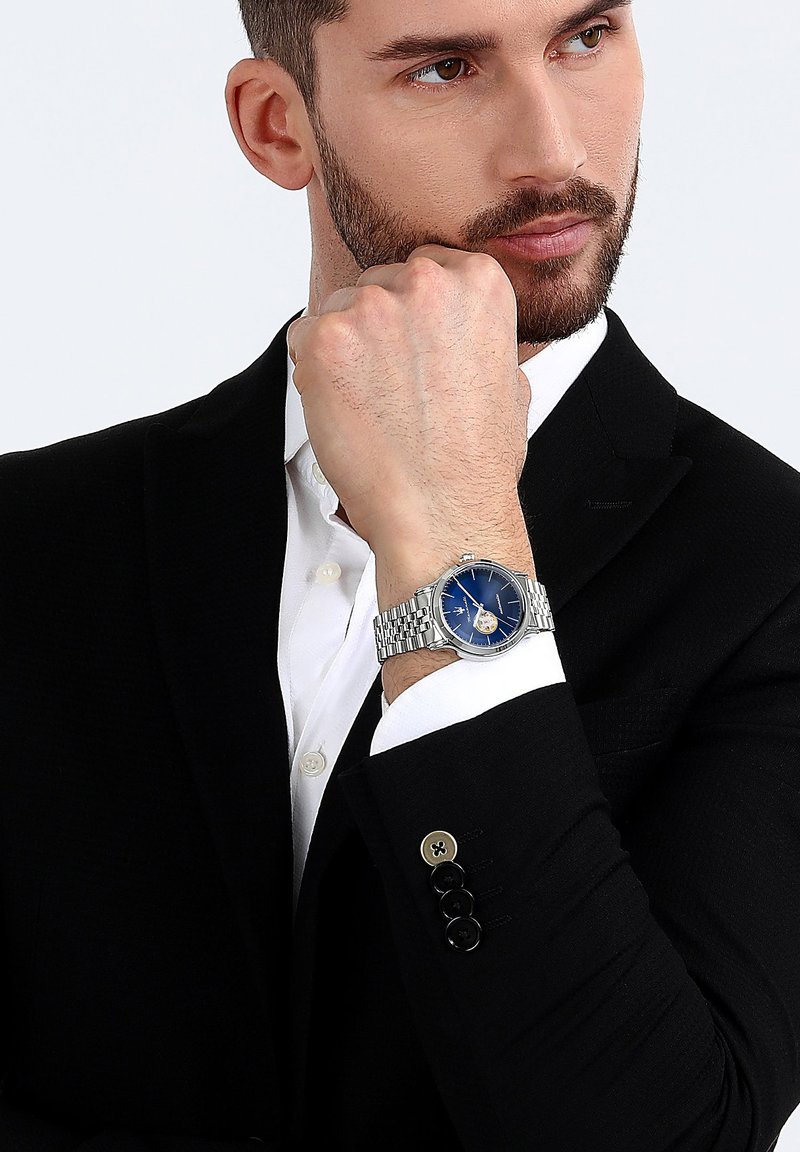 Silver klocka med blå urtavla, armband av rostfritt stål och rund klockkapsel. Klockan har en skeletturtavla och datumvisning.