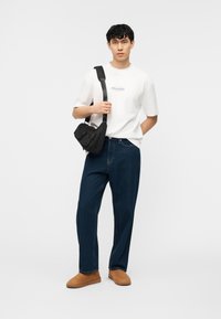 Νέος άντρας με λευκό t-shirt Hollister, σκουρομπλε τζιν, ταμπά παπούτσια slip-on και μαύρη τσάντα χιαστί, με άσπρο φόντο.