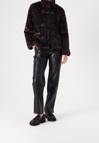 Brązowa kurtka z futerka ekologicznego z zapięciami na troki, zestawiona z czarnymi spodniami z ekologicznej skóry i czarnymi płaskimi butami. Gładka faktura i luźny krój.