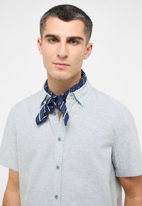Koszula z krótkim rękawem w jasnoszarym kolorze, zapinana na guziki, o teksturowanej tkaninie. Posiada granatowy szalik w biało-skaliste wzory związany wokół szyi.