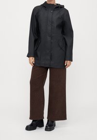 Zwarte waterdichte jas met een capuchon, drukknoopsluiting en voorzakken, gecombineerd met bruine broek met wijde pijpen en een subtiele luipaardprint.