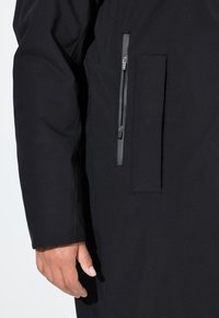 Černé svrchní oděvy se hladkým povrchem mají zapínací kapsu a kapsu na boční švu, navržené pro funkčnost a snadný přístup.