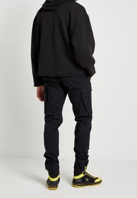 Zwarte cropped hoodie gecombineerd met zwarte cargobroek met zijzakken. Gele high-top sneakers met accenten maken de outfit compleet.