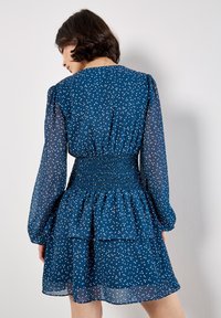 Marineblaues Kleid mit weißen Pünktchen, gerafftem Taillenbund, schichtrock und langen, transparenten Ärmeln. Eleganter, leichter Stoff mit fließendem Design.