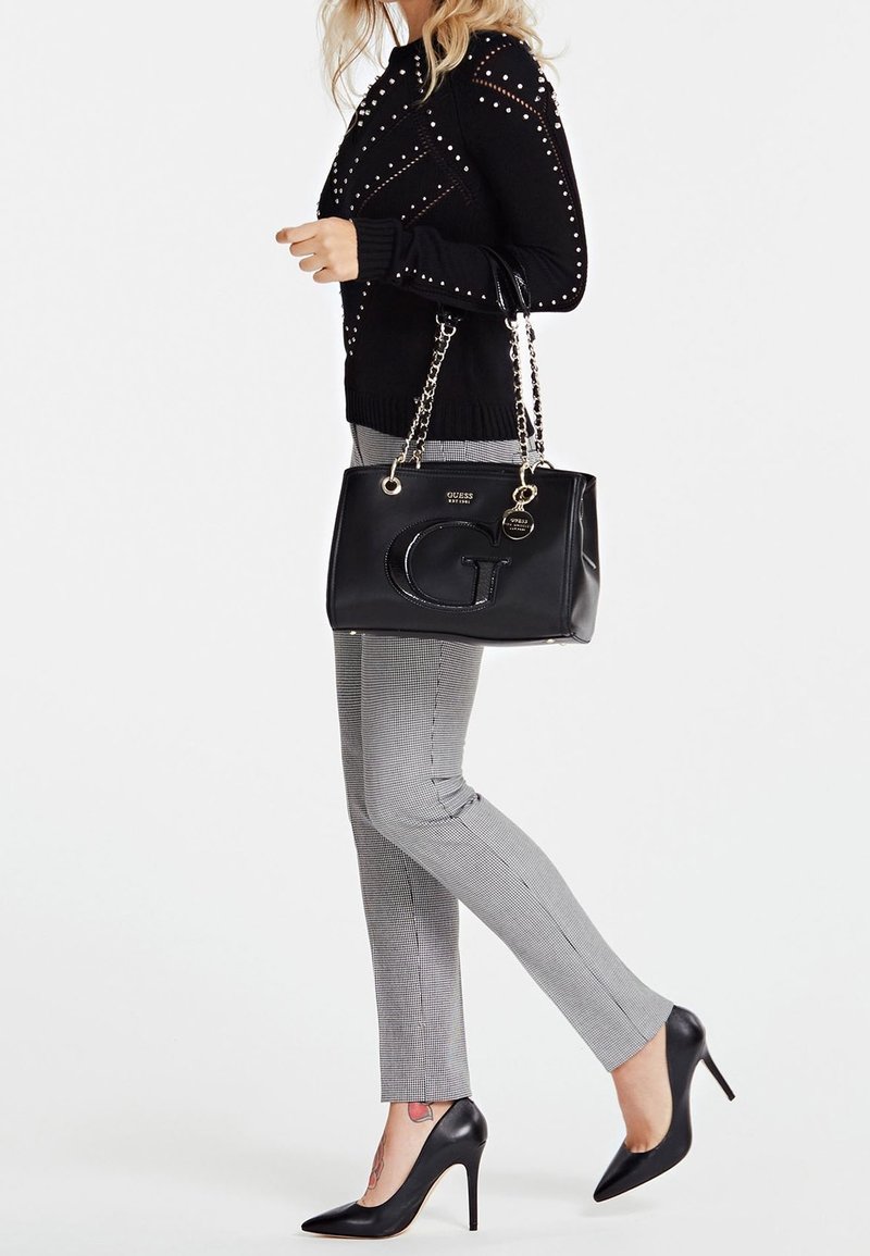 Musta käsilaukku kiiltävällä pinnalla ja ketjusangoilla, yhdistettynä mustaan niittipaitaan, harmaisiin ruutuhousuihin ja mustiin korkokenkiin.