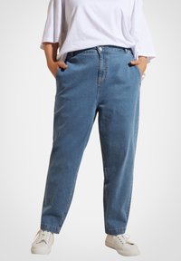 Jeans de mezclilla de color azul claro con corte relajado, que cuentan con bolsillos laterales, cremallera y cierre de botón. Combinados con zapatillas blancas.