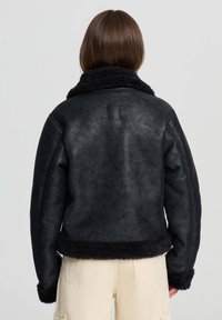 Donna vista di spalle che indossa una giacca in shearling nera con finitura testurizzata e pantaloni crema su uno sfondo chiaro uniforme.
