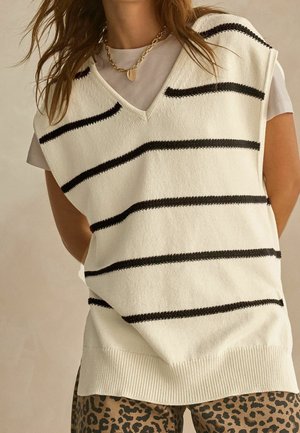 Kobieta w białej dzianinowej kamizelce bez rękawów z czarnymi poziomymi paskami, na beżowej koszulce i spodniach w panterkę.