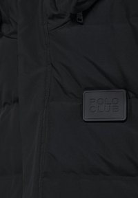 Zwarte gewatteerde jas met een textuur oppervlak, voorzien van een rubberen "POLO CLUB" logo patch op de borst en een gladde stiksel langs de naden.