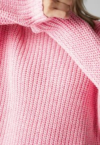 Różowy sweter wykonany na drutach z wąskim splotem i luźnym krojem. Posiada grube mankiety oraz swobodny dekolt, podkreślając przytulny styl.