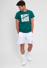 Camiseta verde de manga corta con texto blanco en negrita, combinada con pantalones cortos deportivos blancos. Zapatillas deportivas completan el atuendo.