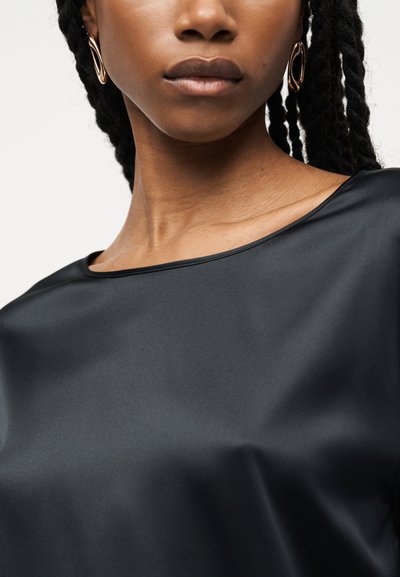 Γυναίκα με σκουρόχρωμα πλεκτά μαλλιά που φοράει χρυσούς στριφτούς κρίκους στα σκουλαρίκια και μαύρο σατινέ τοπ με λαιμόκοψη καραβοκόκαλο, εμφανιζόμενη από τους ώμους μέχρι τα χείλη.