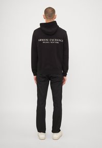 Black hoodie with "ARMANI EXCHANGE MILANO / NEW YORK" printed on the back in white. Soft fabric, standard fit, paired with black pants and white sneakers.