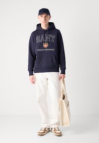 Sudadera de marina con el logo "GANT" en texto blanco, bolsillo frontal, combinada con pantalones blancos y zapatillas marrones. La persona sostiene una bolsa tote beige.