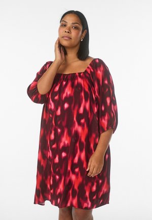 Kvinde med mellemlangt hår iført en løs kjole med rødt og sort mønster, der rører ved sin nakke med den ene hånd.