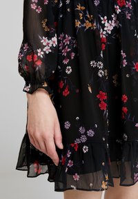 Schwarzes Blumenkleid aus transparentem Stoff, mit langen Puffärmeln und einem rüschengesäumten Saum. Das Design umfasst Blumenmuster in Rot, Lila und Weiß.