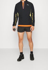 Giacca nera con zip a quarti, dettagli gialli e caratteristiche riflettenti, abbinata a una maglietta arancione e pantaloni corti neri. Tiene in mano una bottiglia d'acqua.