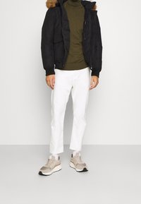 Schwarze Pufferjacke mit einer Kapuze aus Kunstfell, grüner Rollkragenpullover, weiße Slim-Fit-Hose und beiges Sneakers mit grauen Akzenten.