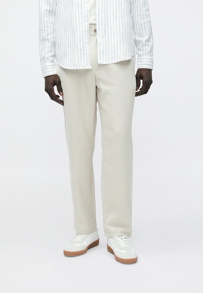 Calças bege claro feitas de tecido macio, com corte reto e textura sutil. Combinadas com ténis brancos com solas de borracha.