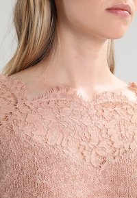 Hellrosa Pullover mit einem Spitzen-Ausschnitt, der mit floralen Mustern verziert ist, aus strukturiertem Strickmaterial und mit ausgefransten Kanten am oberen Rand.