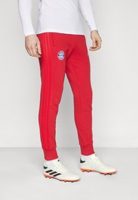 Røde joggebukser med tre striper, utstyrt med Bayern München-logoen. Laget av mykt stoff, smal passform, vist sammen med hvite joggesko.