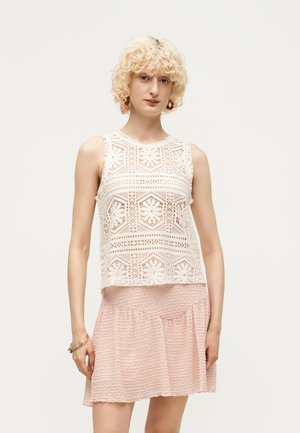 Jeune personne aux cheveux blonds bouclés portant un haut sans manches en dentelle blanche et une jupe rose clair texturée, debout devant un fond uni.