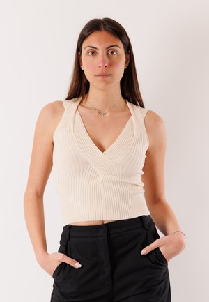 Donna con capelli lunghi e scuri che indossa un top crema a coste senza maniche e pantaloni neri, in piedi con le mani nelle tasche davanti a uno sfondo semplice.