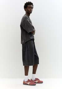 T-shirt oversize grigio scuro con maniche corte, abbinato a pantaloni corti fino al ginocchio. Il modello indossa scarpe da ginnastica rosse e bianche con calze bianche.