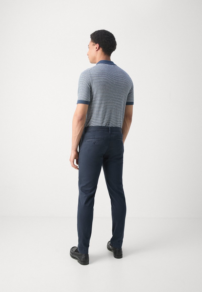 Herren Polo-Shirt mit kurzen Ärmeln in Blau- und Weißstreifen, kombiniert mit marineblauen Hosen. Das Polo verfügt über einen kontrastierenden Kragen und einen strukturierten Strickstoff.