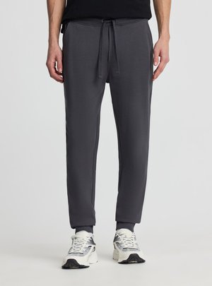 Uomo con pantaloni jogger grigio scuro a gamba stretta e sneakers bianche, in piedi davanti a uno sfondo chiaro e semplice.