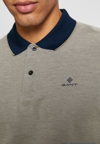 Koszulka polo w odcieniu szarym z teksturowanym wzorem, granatowym kołnierzykiem i subtelnymi zielonymi akcentami. Posiada zapięcie na dwa guziki oraz logo GANT na klatce piersiowej.