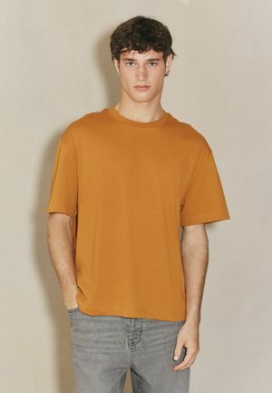 T-Shirt mit kurzen Ärmeln und Rundhalsausschnitt in einem warmen Orangeton, aus weichem Stoff gefertigt, kombiniert mit hellgrauen Jeans. Minimalistisches Design, lässige Passform.