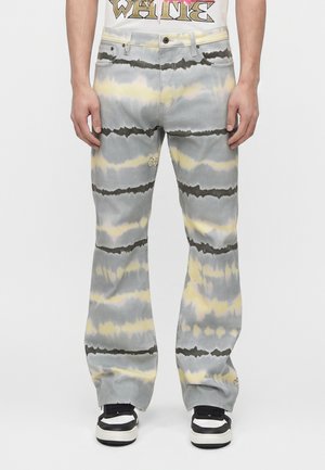 Mężczyzna w szarych, żółtych i czarnych spodniach tie-dye w paski oraz czarno-białych trampkach, stojący na jednolitym tle.
