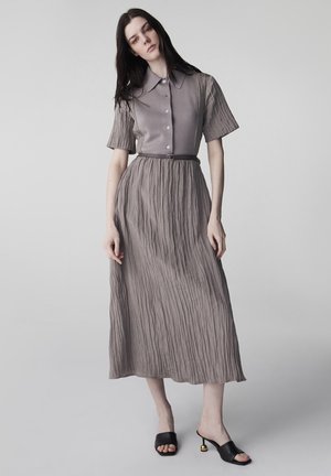 Kobieta ubrana w teksturowaną, szarobrązową sukienkę midi z krótkimi rękawami i kołnierzem, w połączeniu z czarnymi, odkrytymi na palce sandałami na obcasie ze złotymi akcentami.