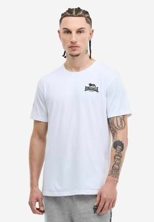Jeune homme aux cheveux tressés portant un t-shirt blanc Lonsdale London et un short gris, exhibant des tatouages détaillés sur son bras droit.