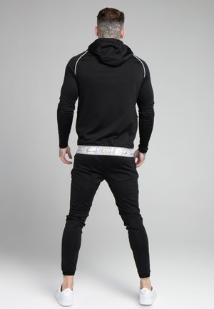 Homme avec tatouages sur le cou et les mains, portant un sweat à capuche noir et un pantalon avec des baskets blanches, debout face à un fond gris, dos tourné.