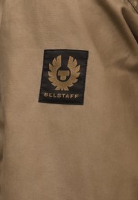 Μαύρο ορθογώνιο έμβλημα λογότυπου Belstaff με χρυσό σήμα και κείμενο σε ταμπά υφασμάτινο φόντο.