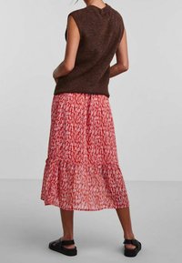 Brun ärmlös stickad topp ihop med en röd lagerkjol med vita abstrakta mönster. Modell som bär svarta sandaler.