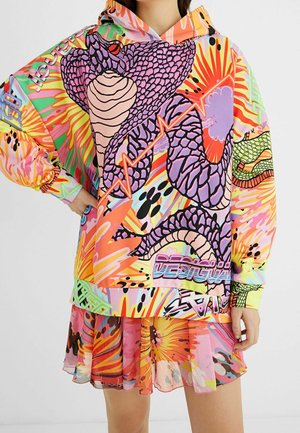 Sweat à capuche oversize coloré et jupe assortie avec des motifs serpent et abstraits vibrants porté par une personne avec la main sur la hanche.