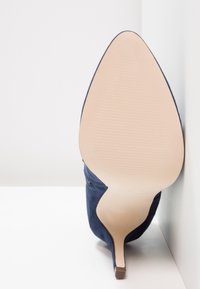 Granatowe zamszowe buty na wysokim obcasie z ostrym noskiem i smukłym obcasem typu stiletto. Podeszwa jest gładka i beżowa z delikatną teksturą.
