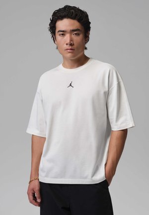 Ung mand med krøllet sort hår iført en hvid Jordan t-shirt og sorte bukser, stående foran en ensfarvet grå baggrund.