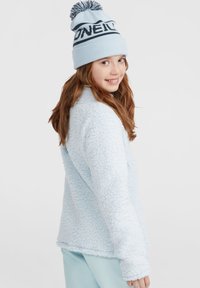 Veste en polaire bleu clair avec une surface texturée et duveteuse, associée à un bonnet en tricot bleu clair avec une bande plus foncée et un pompon.