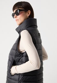 Doudoune noire sans manches avec un col haut, design matelassé, poche latérale et une silhouette épurée, portée sur un haut à côtes léger.