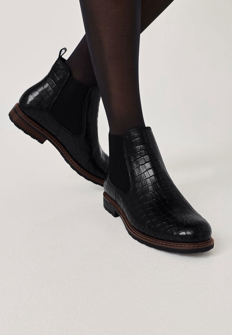 Bottines en cuir noir à texture croco avec panneaux élastiques sur les côtés, portées avec des collants noirs transparents, présentées sur un fond clair.
