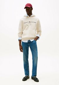 Felpa beige con logo "TOMMY HILFIGER", camicia azzurra sotto, jeans blu e scarpe nere. Un cappellino rosso completa il look.