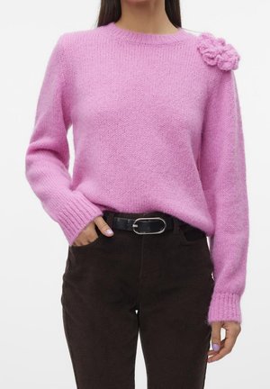Femme portant un pull en maille rose avec un détail fleur sur l'épaule, ceinture noire et pantalon en velours côtelé brun foncé.