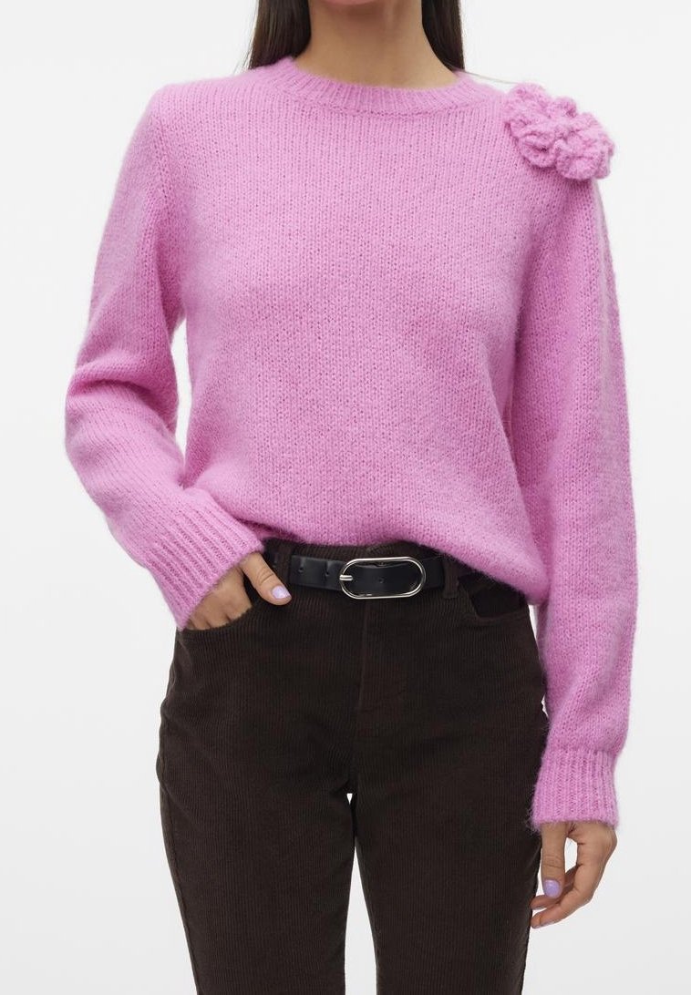 Femme portant un pull en maille rose avec un détail fleur sur l'épaule, ceinture noire et pantalon en velours côtelé brun foncé.