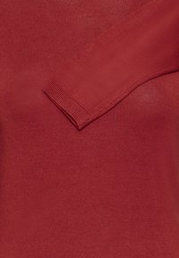 Karmī sarkans adīts džemperis ar ribotu tekstūru un plašu apkakli. Piedurkne ir daļēji redzama, parādot bezšuvju dizainu.