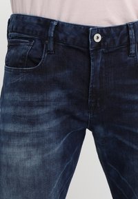 Jeans de mezclilla azul oscuro con un lavado ligeramente descolorido. Incluye un bolsillo frontal, cierre de botón y trabillas para el cinturón.