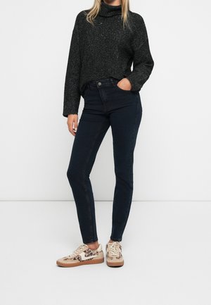 Femme portant un pull à col roulé gris foncé, un jean skinny foncé et des baskets beige à motif léopard, debout avec une main dans la poche.
